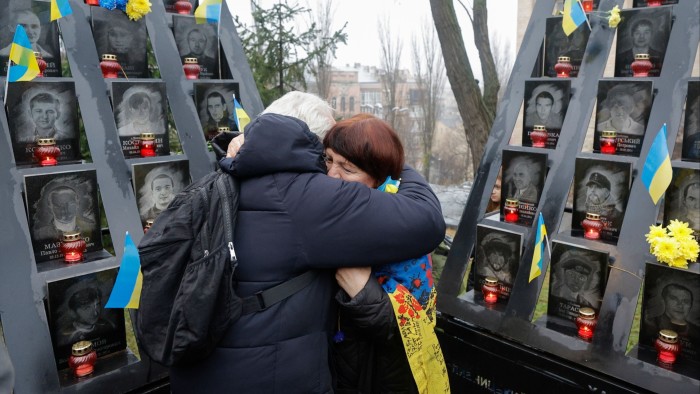 Two people embrace in front of memorial plaques and candles honoring the 'Heavenly Hundred' killed during the Euromaidan protests.