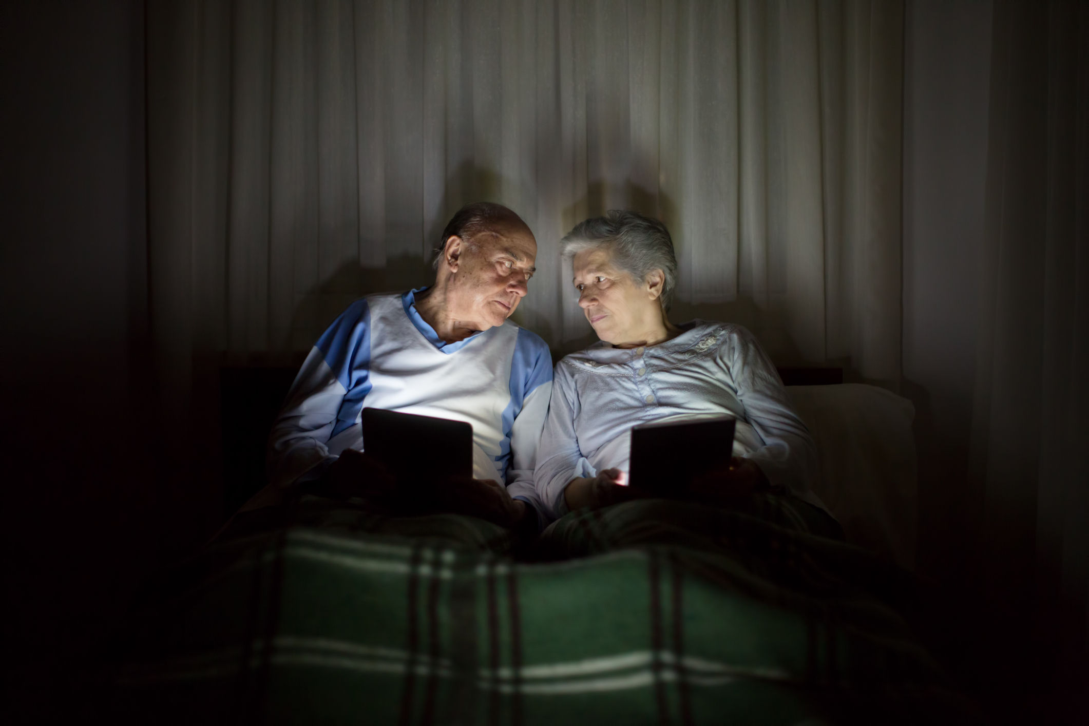 An elderly couple sits in bed, illuminated by the soft glow of their devices. They share a moment of connection and intimacy, engrossed in their screens.