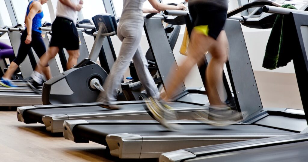 Motion-blurred legs of people walking and running on treadmills in a bright gym. This image illustrates the common New Year's resolution of improving physical fitness, while serving as a visual hook for discussing the financial costs of gym memberships and offering budget-friendly alternatives for 2026 health goals. Source: The College Investor