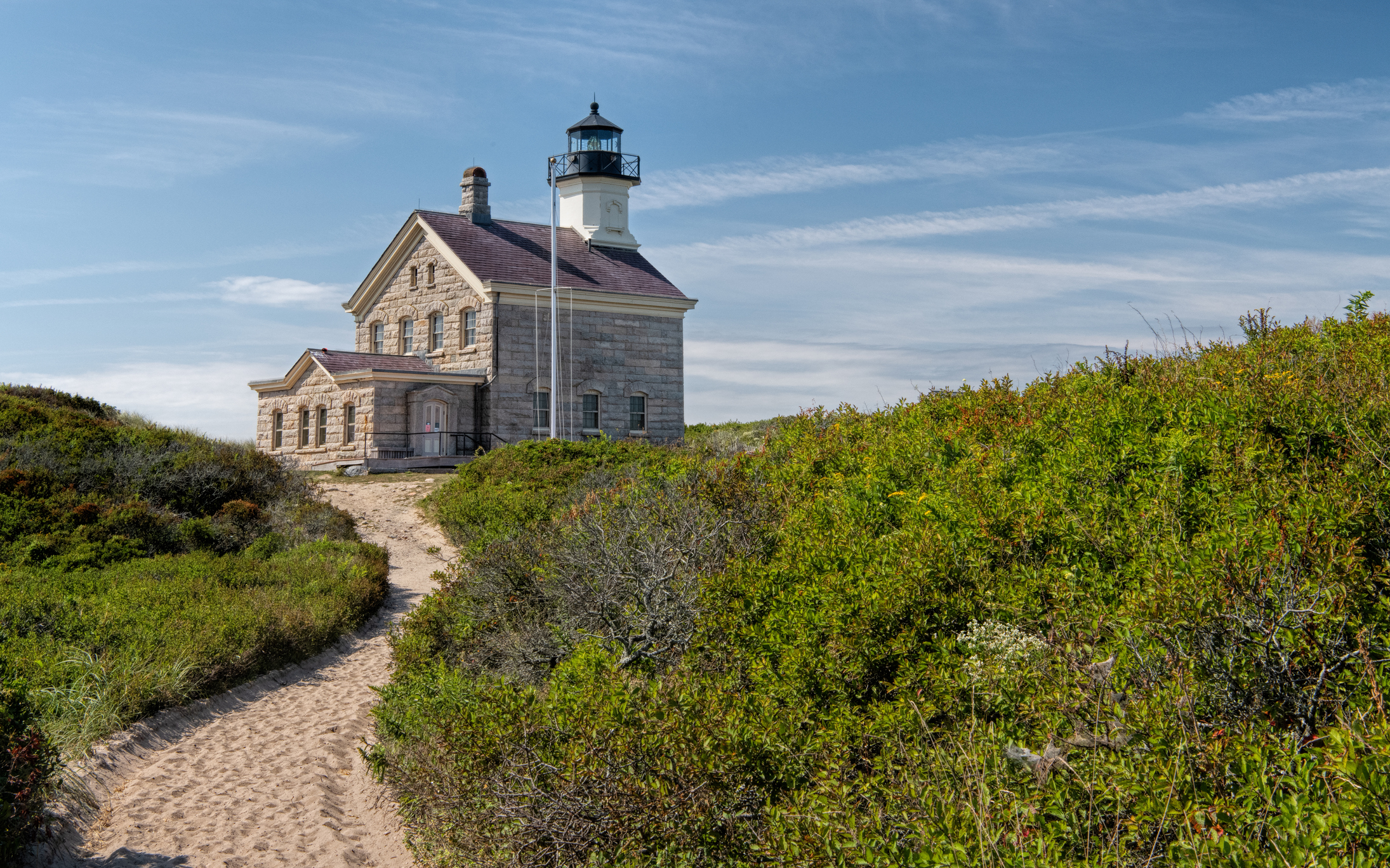 Trail approaching the North Lighthouse on Block Island Sunny day w blue sky Lighthouse is in the center of the landscape surrounded by beach grass and dunes Trail leads a short distance directly to the lighthouse