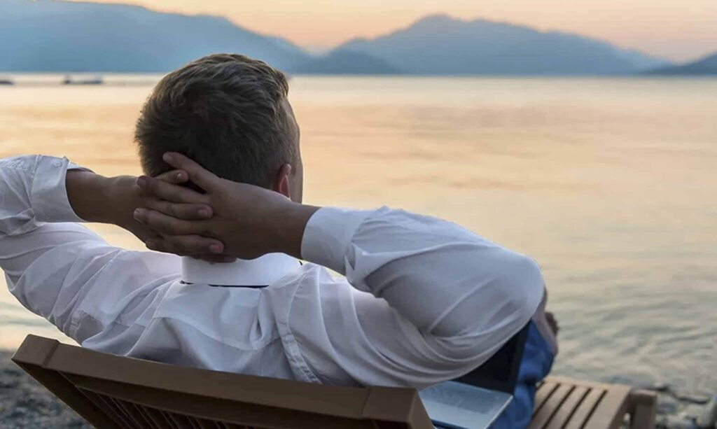 A man with blonde hair, dressed in a white collared shirt, sits relaxed on a wooden bench with his hands clasped behind his head, gazing out over a calm body of water at sunset. A silver laptop is visible next to him on the bench, suggesting he has just finished working or is taking a break while his investments generate passive income. The warm, soft light of the setting sun creates a tranquil atmosphere, emphasizing the freedom and peace of mind associated with achieving financial independence through passive income streams. This image perfectly illustrates the article's core message about earning money without continuous active effort, highlighting the desired outcome of strategic monetary or time investments. Source: The College Investor