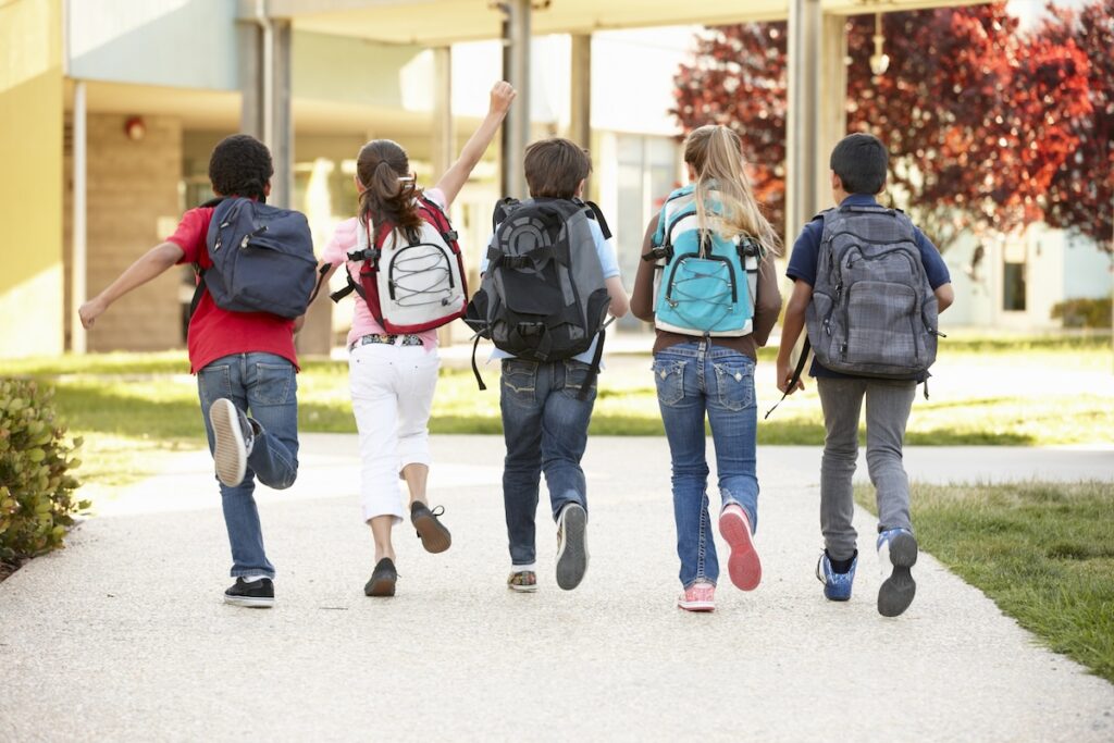 Diverse group of smiling elementary school children running away from the camera down a sunny walkway lined with trees and school buildings. The children are wearing backpacks, representing the broad range of K-12 students who could benefit from the new Education Freedom Tax Credit, which aims to fund scholarships for public, private, and charter school expenses. Source: The College Investor