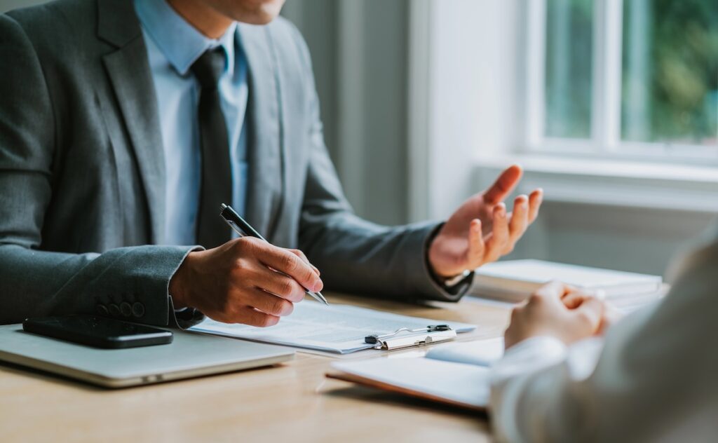 Close-up of a businessman in a gray suit holding a pen while explaining a contract to a client at a wooden table. This image illustrates the consultation process for refinancing private student loans, specifically highlighting the new option of income-based repayment plans offered by lenders like RISLA. Source: The College Investor