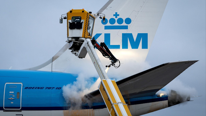 A KLM Boeing 787-10 plane being sprayed with de-icing fluid by a yellow de-icing vehicle at Schiphol Airport.