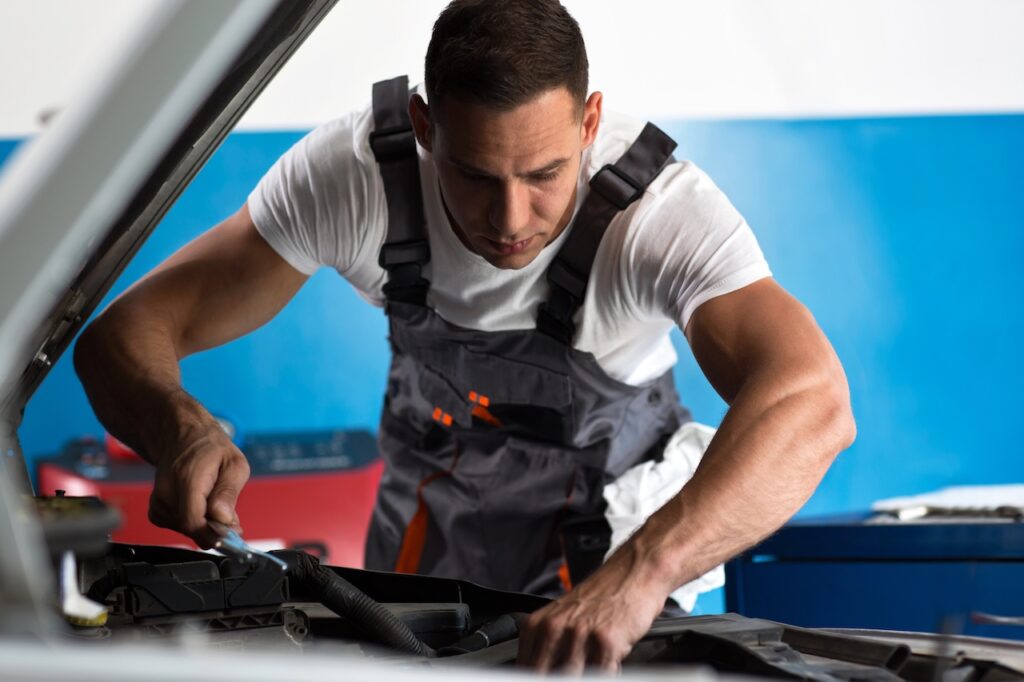 Close-up of a focused mechanic wearing a white T-shirt and grey overalls working under the open hood of a car, holding a wrench. This image serves as a visual metaphor for the student loan rehabilitation process&mdash;a deliberate, hands-on repair strategy that allows borrowers to fix their defaulted loans and restore their financial standing through consistent effort. Source: The College Investor