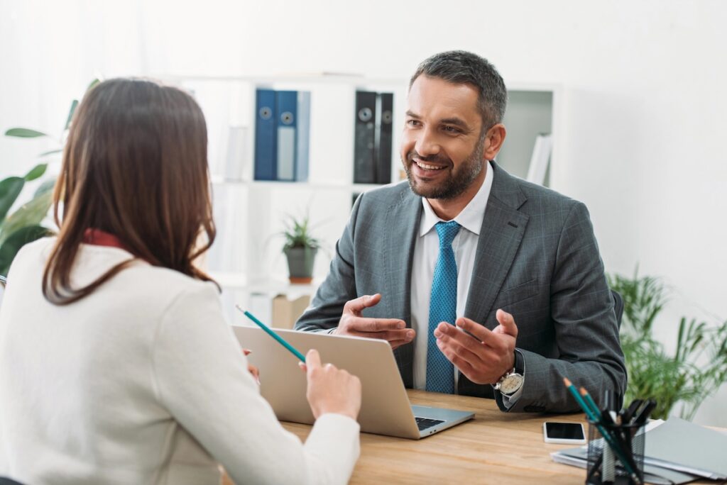 A smiling businessman in a grey suit and blue tie gestures while speaking to a client in a bright office. This image represents the role of financial professionals&mdash;specifically Certified Financial Planners (CFPs) or student loan consultants&mdash;who provide strategic advice to borrowers navigating complex repayment options, forgiveness plans, and long-term financial goals. Source: The College Investor