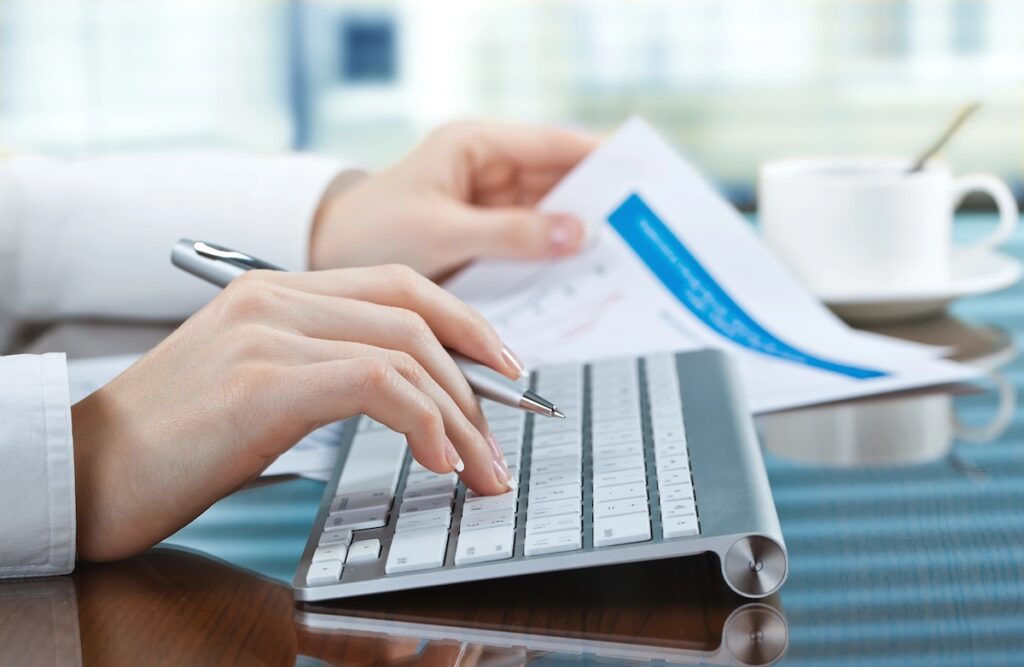 Close-up of a person&rsquo;s hands typing on a modern keyboard next to financial documents and a cup of coffee. This image illustrates the process of managing tax information, specifically focusing on Form 1098-T, scholarships, and understanding the tax implications for students and families. Source: The College Investor