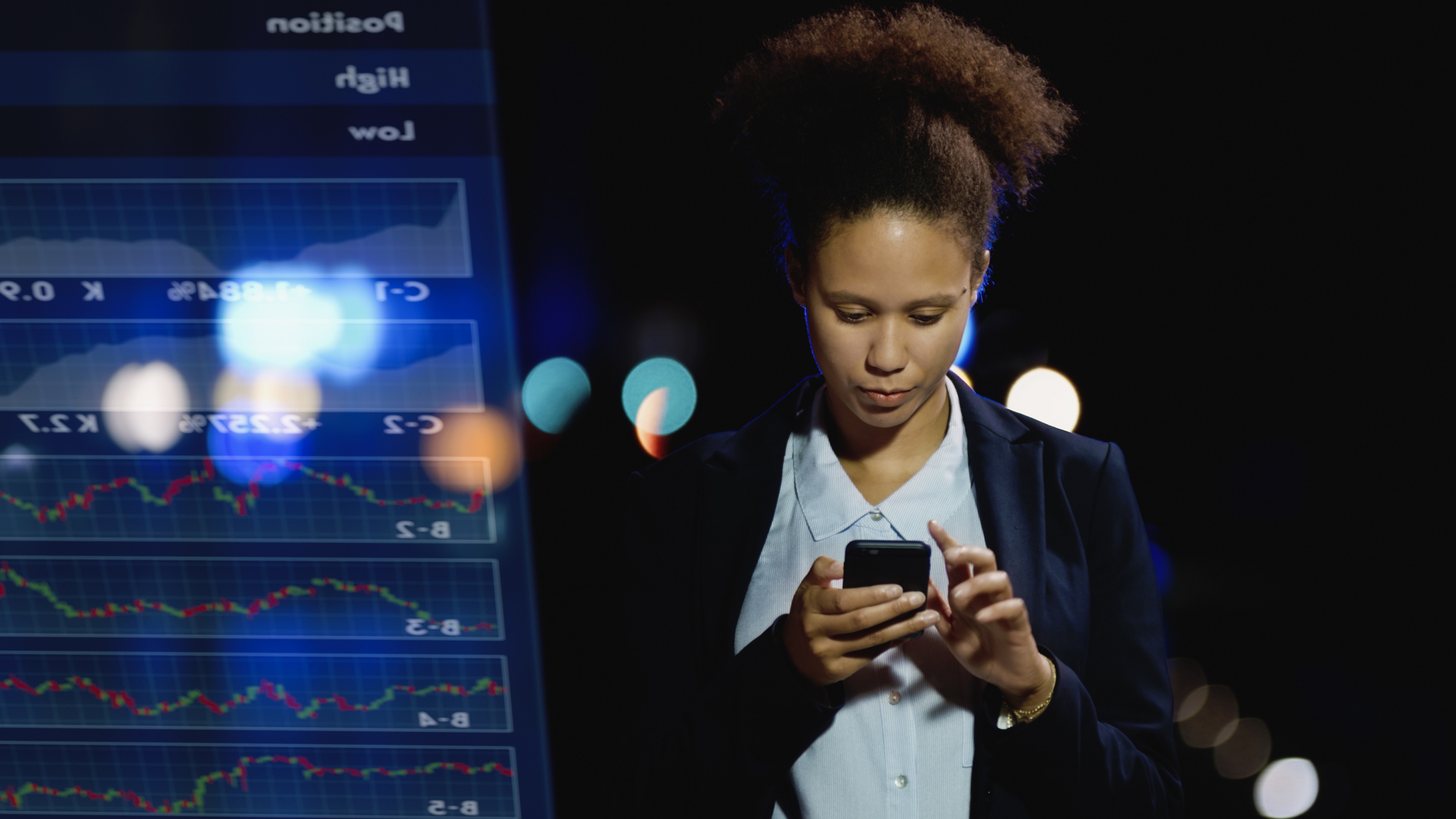 A woman looks at her phone while standing next to a trading monitor.