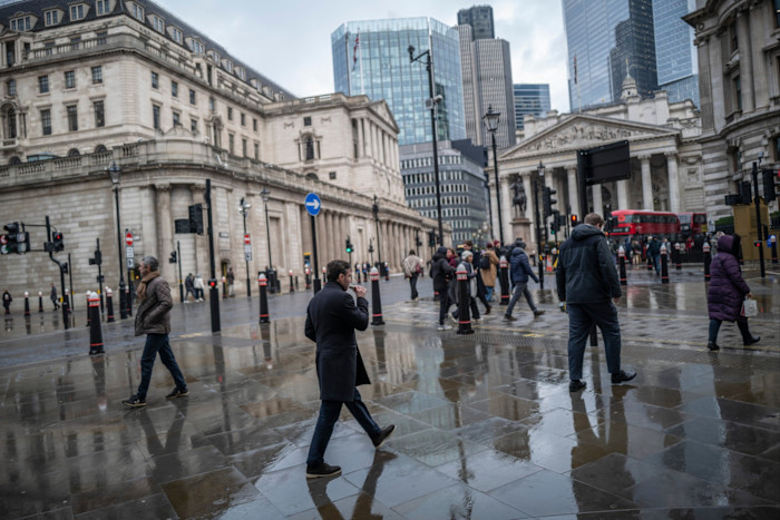 Pedestrians walk across wet pavement outside the Bank of England and the Royal Exchange, with city buildings and a red double-decker bus in the background.