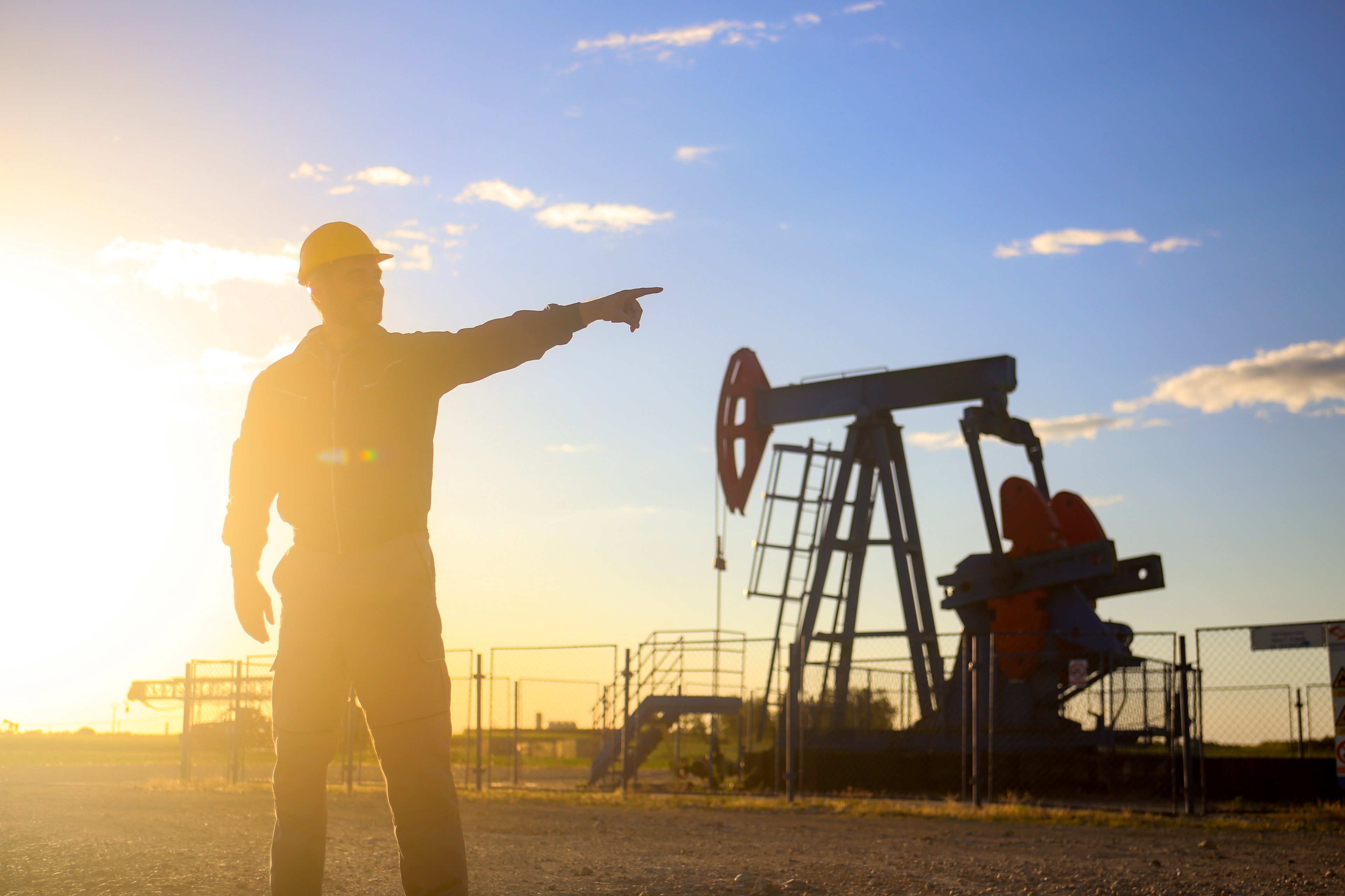 An oil field worker wearing a hard hat and pointing to an operating pumpjack in the distance.
