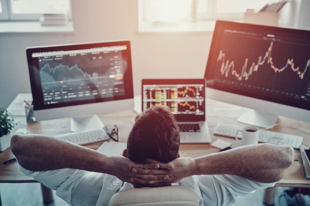 A person leans back in a desk chair with their hands behind their head while facing several computer monitors displaying data.