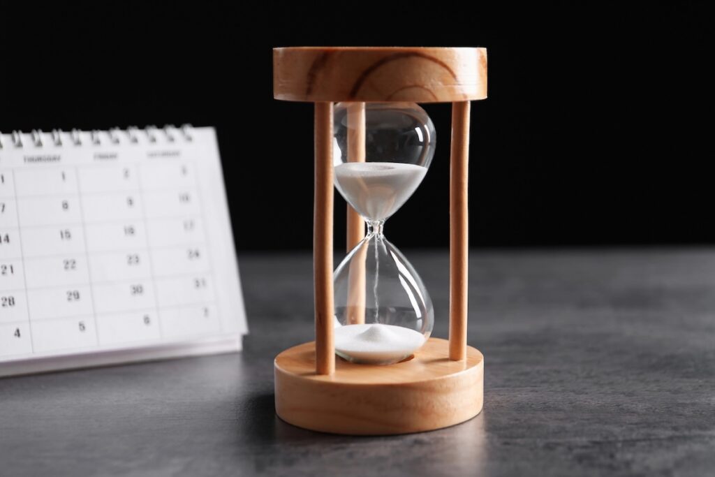 Close-up of a wooden hourglass with sand running out, sitting next to a white paper calendar on a dark surface. This image visualizes the urgency of the upcoming July 1, 2026, deadline for federal student loan borrowers&mdash;especially Parent PLUS holders&mdash;to consolidate their loans, or risk permanently losing access to income-driven repayment plans and forgiveness programs. Source: The College Investor