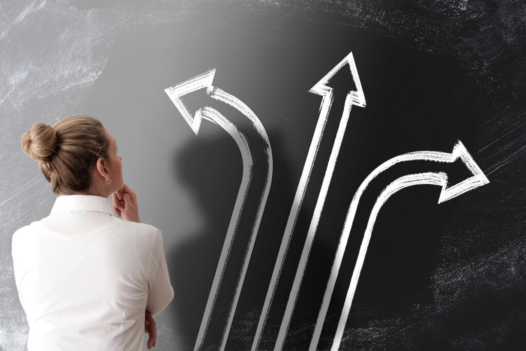 Rear view of a businesswoman in a white shirt looking up at a chalkboard with three large arrows pointing in different directions. This conceptual image illustrates the complex decision-making process parents face regarding student loans in 2026, as new federal limits on Parent PLUS loans force families to weigh federal versus private borrowing options. Source: The College Investor