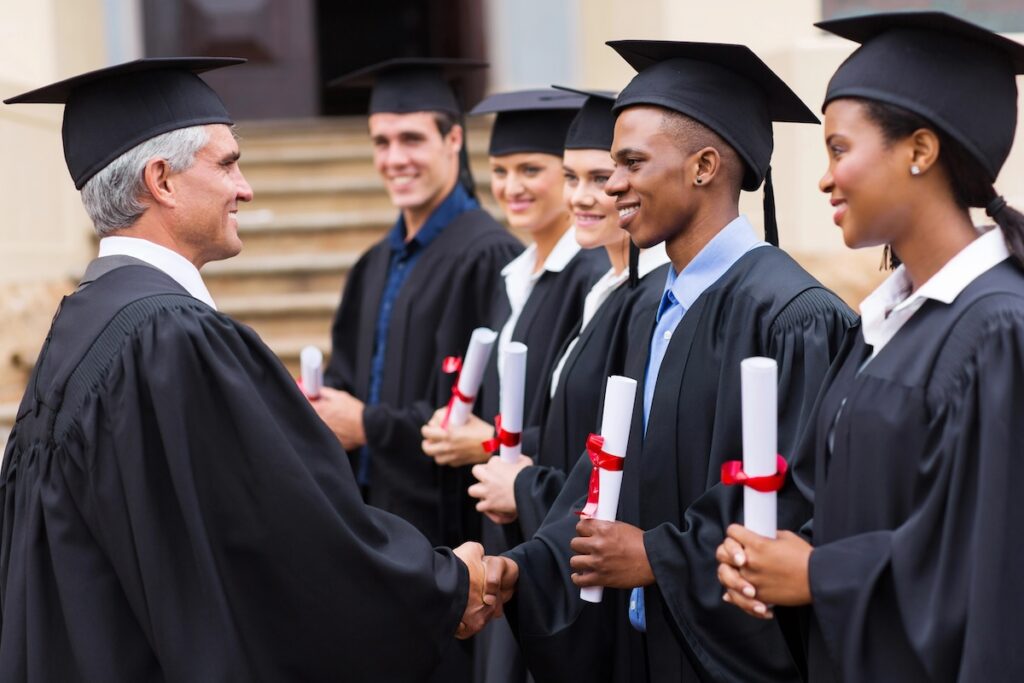 Side view of a diverse line of college graduates in black caps and gowns shaking hands with an administrator and receiving their diplomas. This image represents the beginning of the post-graduation journey, where nearly one in three borrowers report that student loan debt is delaying major life milestones like homeownership and retirement savings. Source: The College Investor