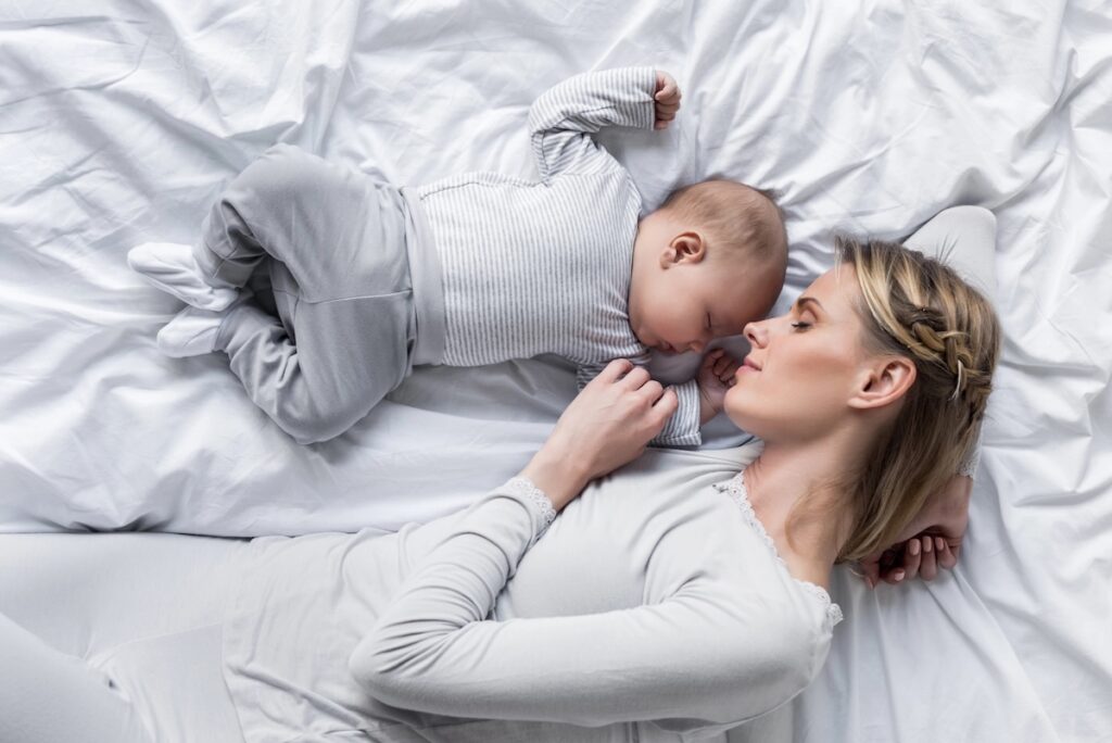 Top-down view of a mother wearing white pajamas sleeping peacefully next to her newborn baby on a white bed. This image accompanies a guide on navigating student loan repayment during maternity leave, illustrating how mothers can manage reduced income and rising household expenses through deferment, forbearance, or income-driven repayment strategies. Source: The College Investor