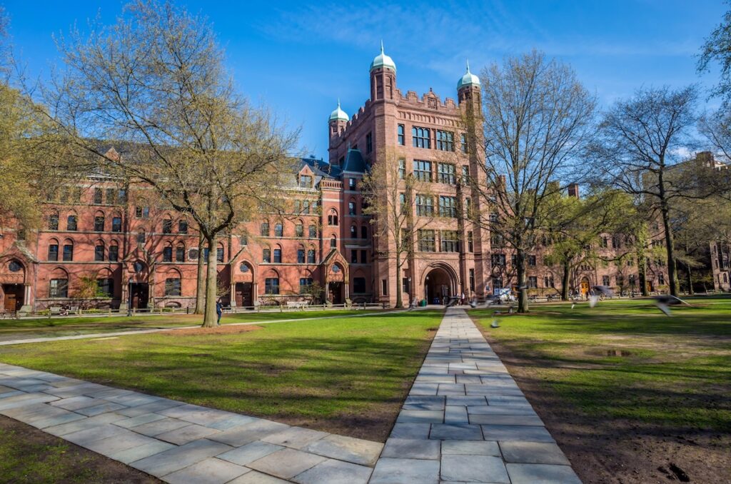 Sunny view of a paved pathway leading toward the historic, castle-like red brick buildings of Yale University's campus. This image visually anchors the news of Yale raising its free-tuition income threshold to $200,000, highlighting the elite institution's efforts to expand affordability amidst rising costs. Source: The College Investor