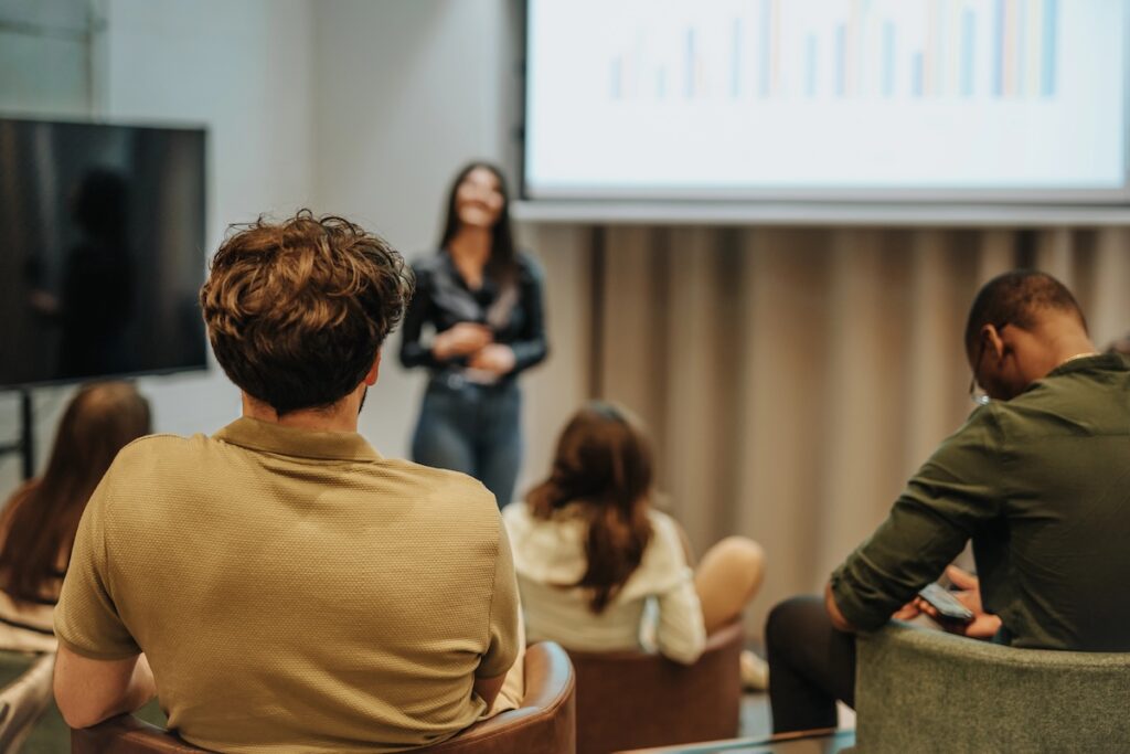 Rear view of a diverse group of adults sitting in a modern classroom, looking at a large screen displaying a colorful bar chart. A woman stands presenting, while attendees in casual attire focus on the data. This image illustrates the