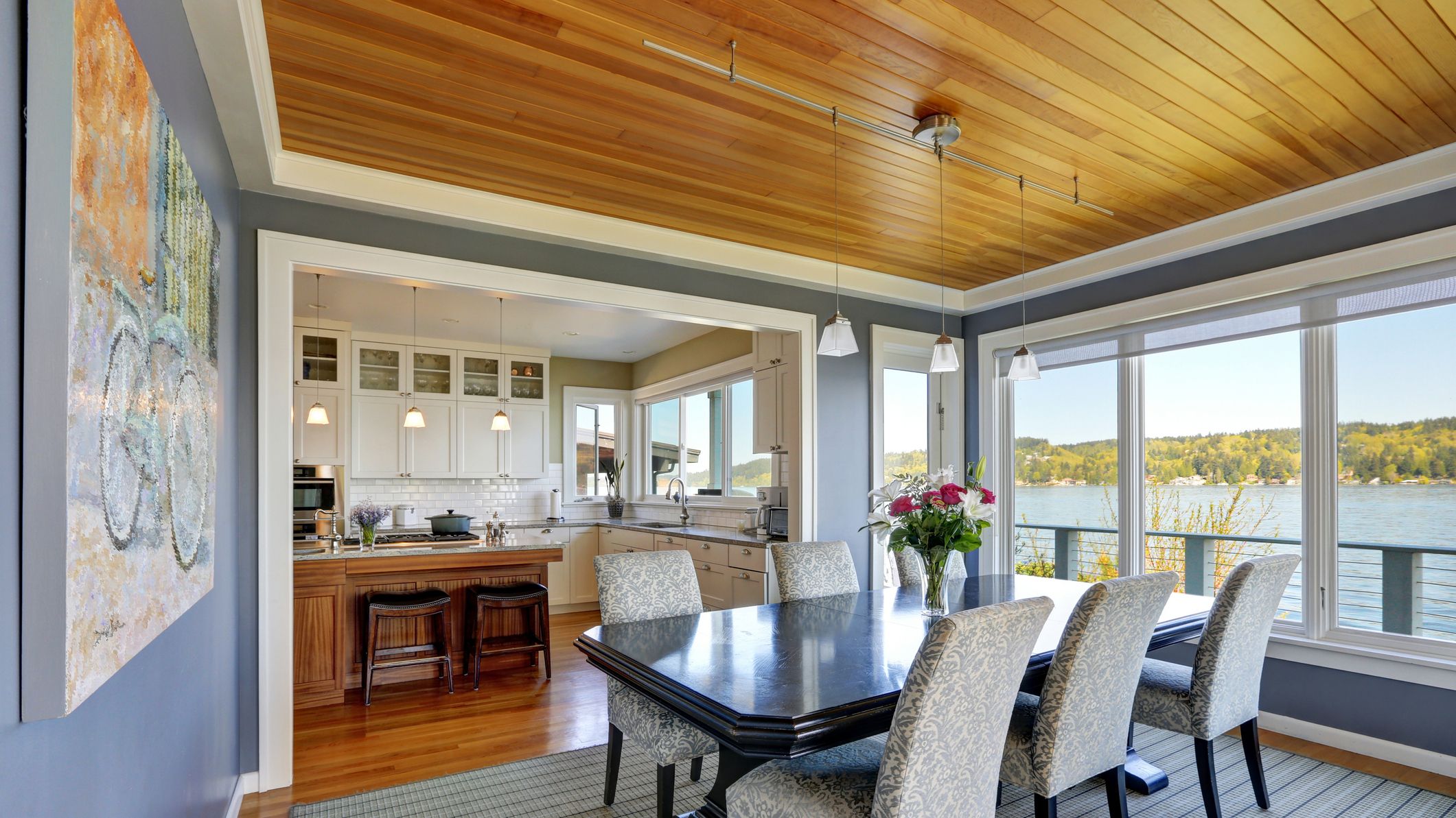 Formal dining room features navy blue walls, wood plank ceiling over black dining table with floral parsons chairs atop grey striped rug. Large windows overlook Lake Washington. Northwest, USA.