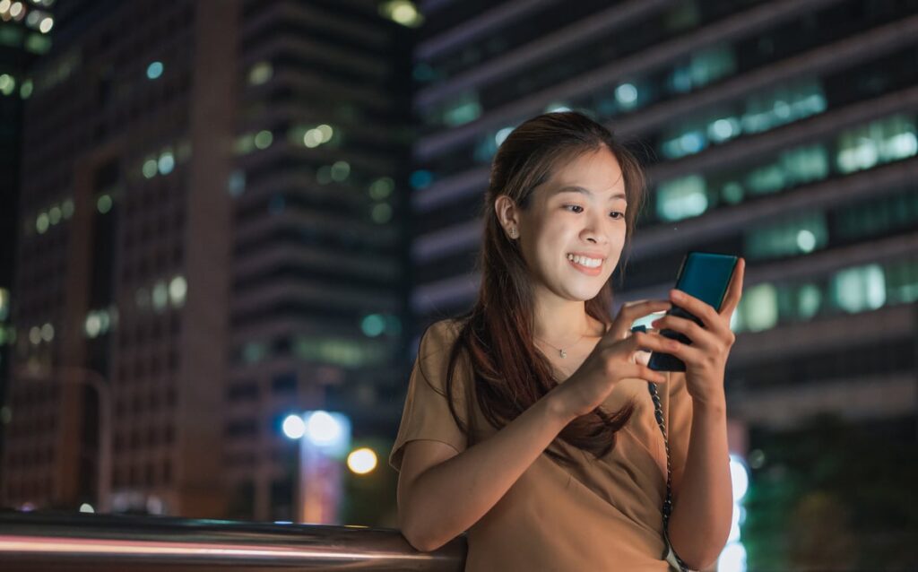 A person smiling, looking at phone, with the night-time city landscape behind them.