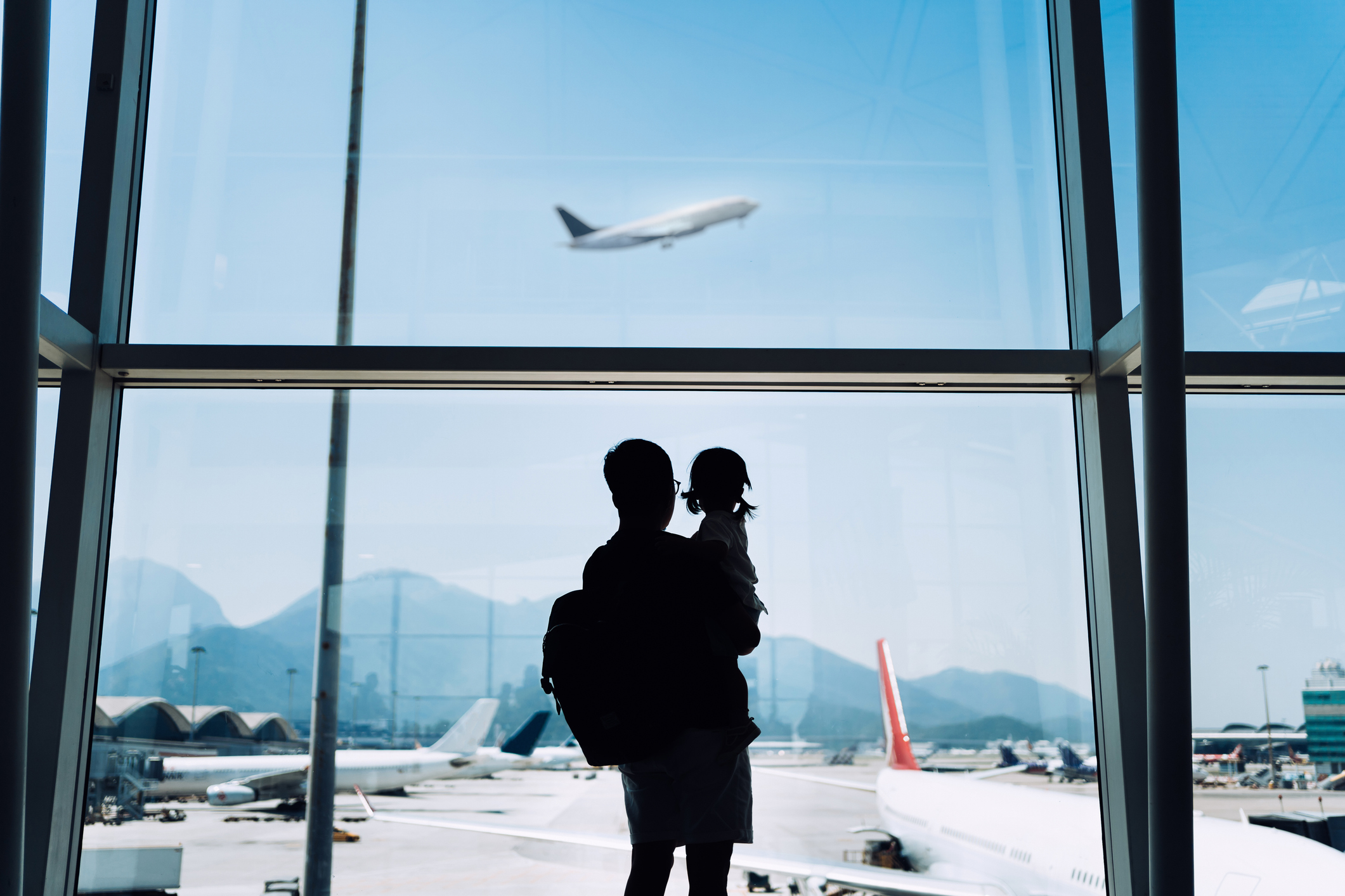 A man holds a little girl up to look at a plane taking off through windows at an airport.