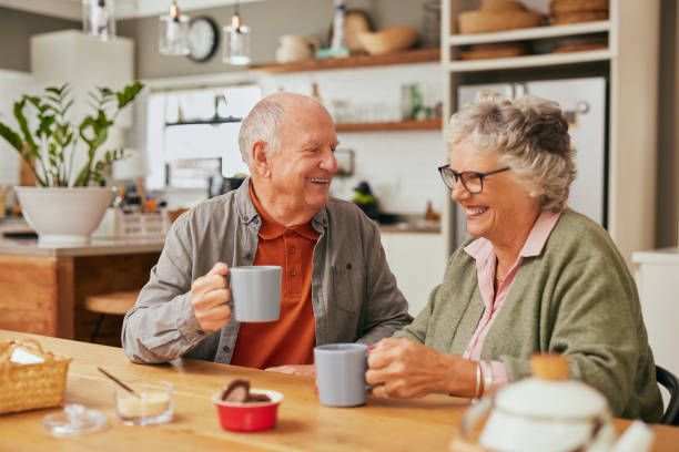 Smiling old couple sitting at table and sharing coffee in their cozy kitchen. Elder man and retired woman laughing together while enjoying a cup of coffee indoors.