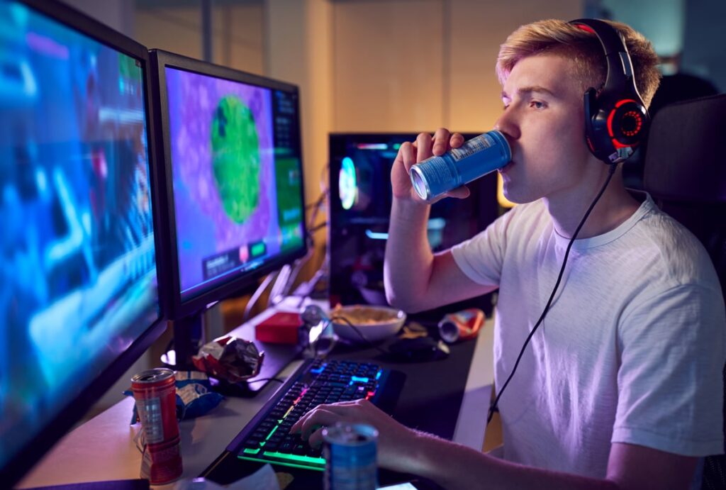 A gamer drinking an energy drink while playing video games with two side-by-side monitors.