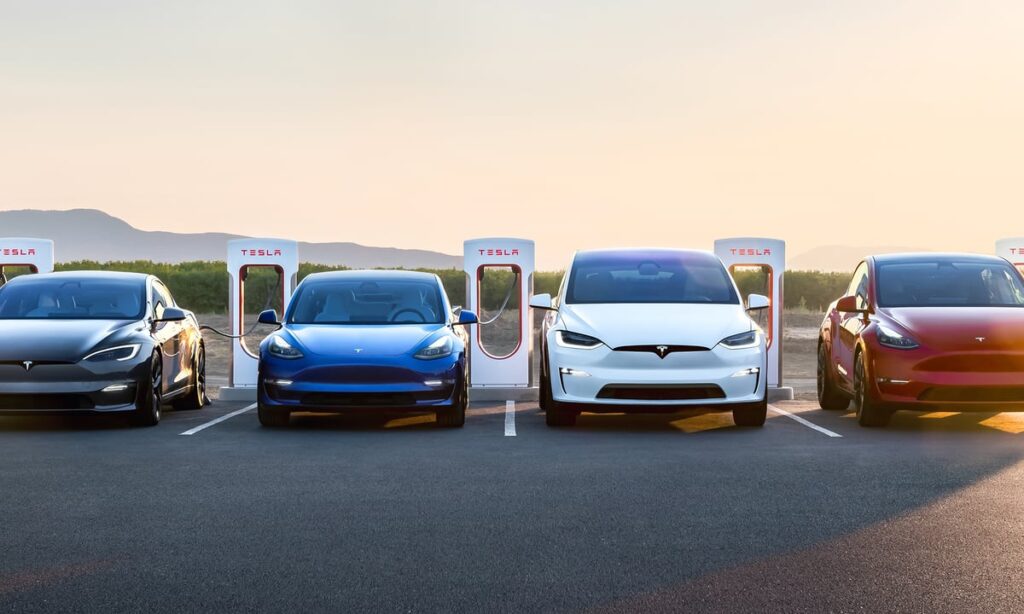 Four Tesla cars parked at charging stations with low-lying hills in the background.