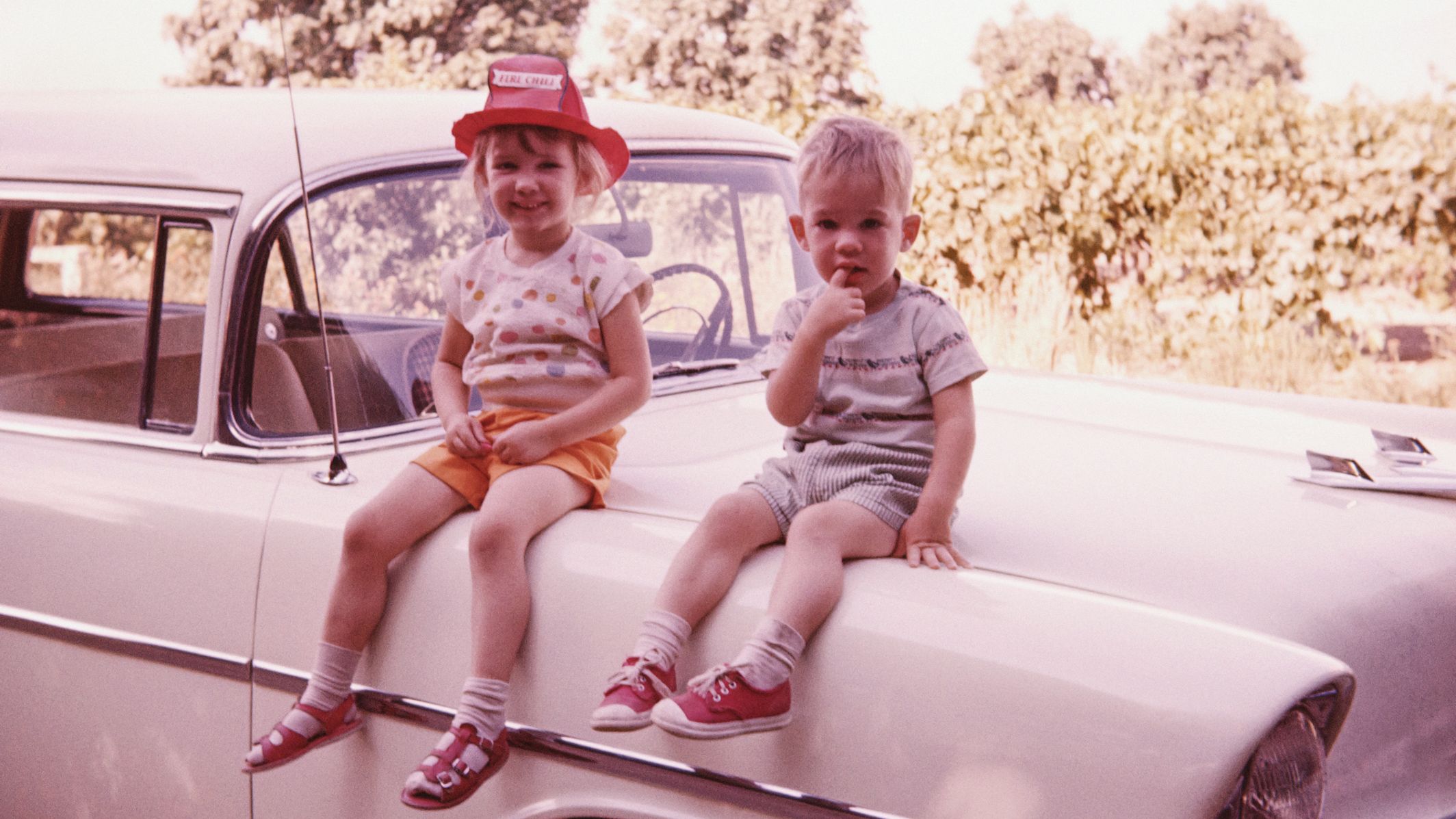 A vintage 1950s photograph of a brother and sister sitting on the hood of a car.