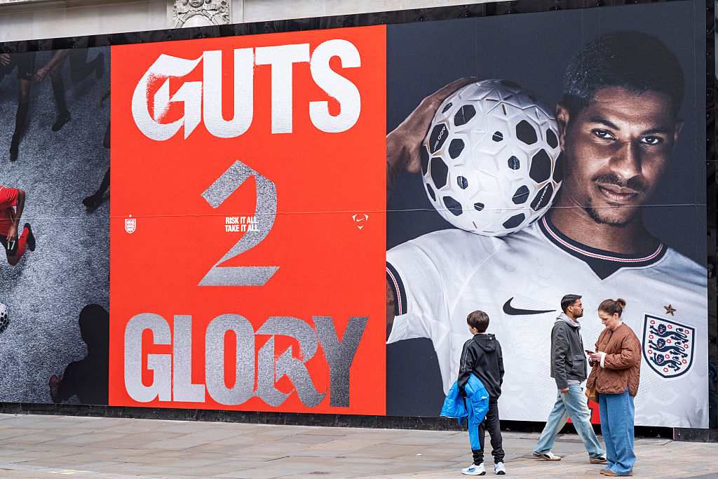 People standing beside a World Cup mural that reads "Guts 2 Glory" and has a soccer player in a Nike jersey