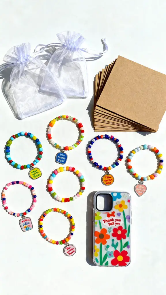 A flat-lay display of handmade beaded bracelets and phone charms next to packaging bags and thank-you cards on a white surface.