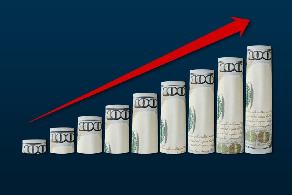 Rising rolls of $100 bills with a rising red arrow.