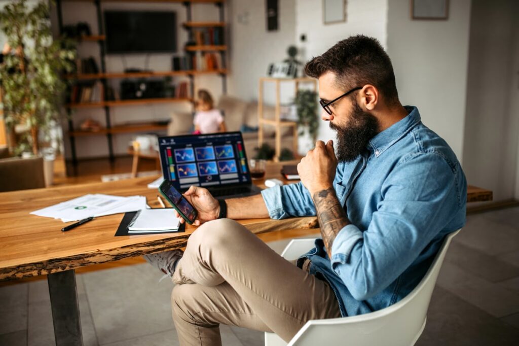 Person at desk comparing investments on cellphone and computer.