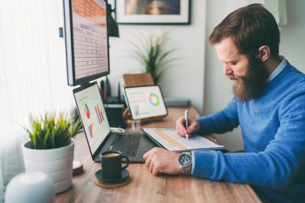Person writing note in notebook and looking at stock charts.
