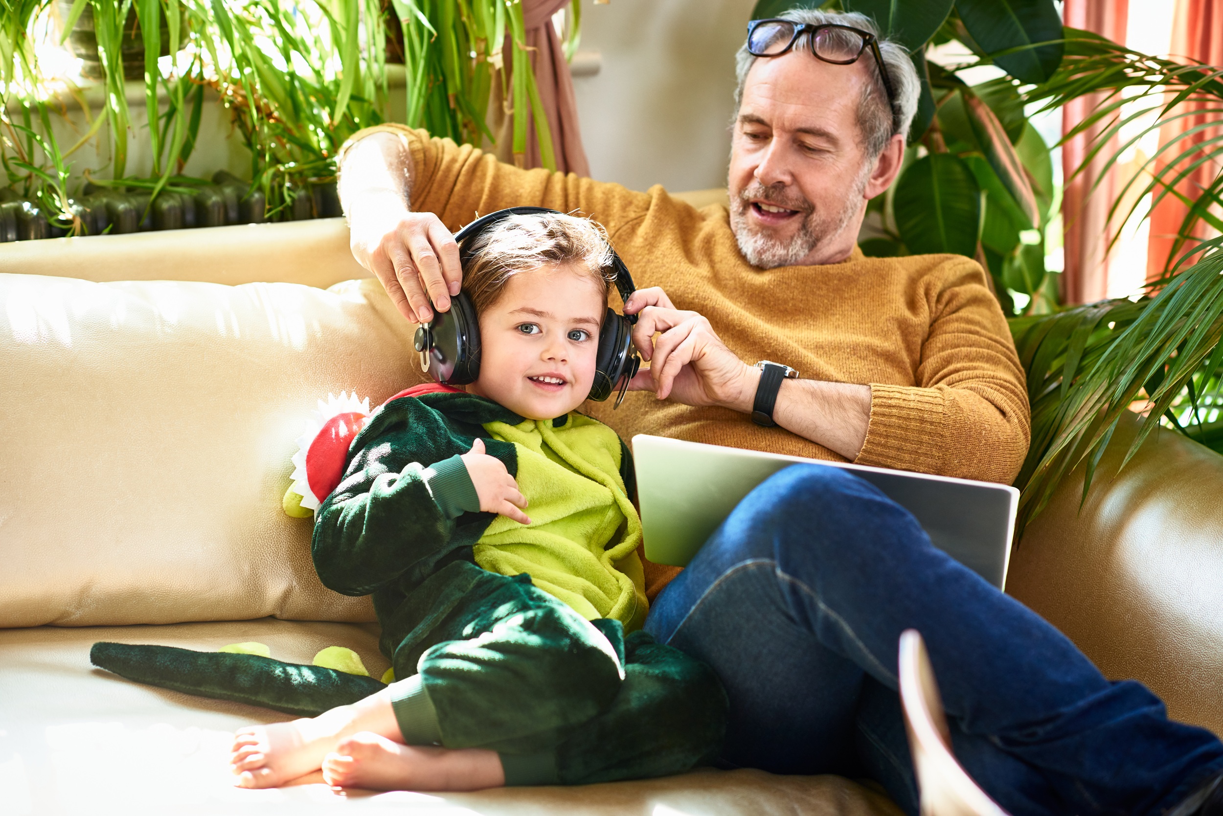 Cute child wearing dinosaur outfit listening to music, sitting on sofa with grandfather, discovery, sensory perception, development