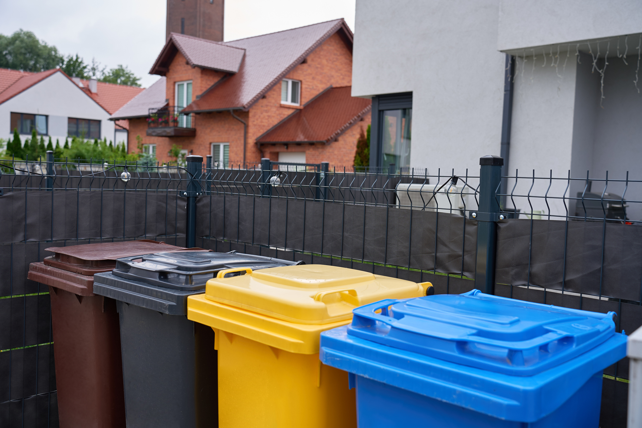 Colored recycling bins stand behind a fence in a residential neighbourhood near modern houses.