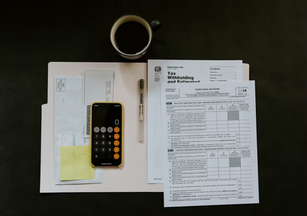 IRS tax forms, a pen and a calculator app on a phone laid out on a folder beside a cup of coffee