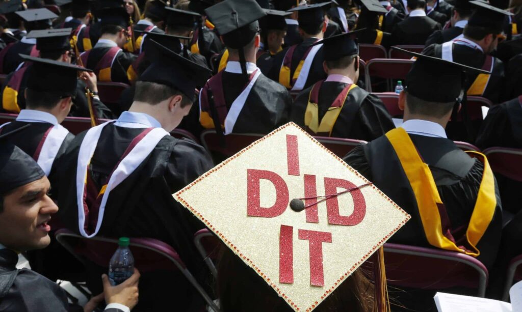 Rear view of a crowd of college graduates in black gowns and maroon-and-gold hoods sitting in rows during a commencement ceremony. In the center focus, a student wears a decorated mortarboard covered in gold glitter with large red sparkling letters spelling