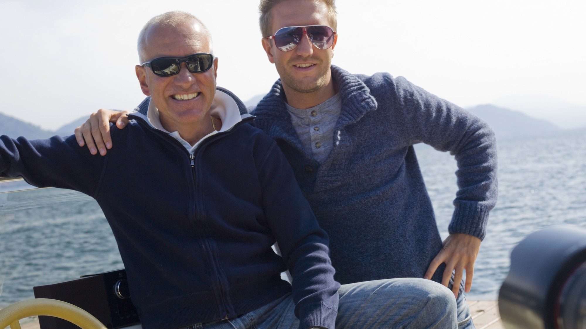 A mature father and his adult son smile from a sail or motor boat.