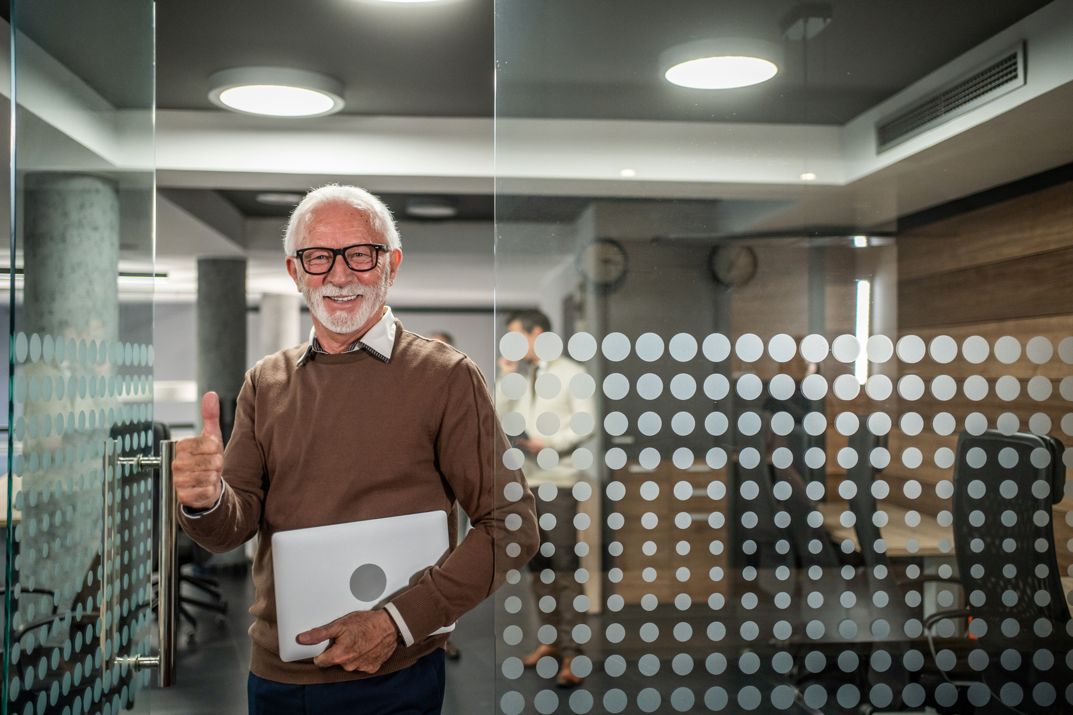 Senior professional man with a laptop standing in a contemporary office, confidently smiling and giving a thumbs up gesture, symbolizing success, expertise, and positive business outcomes