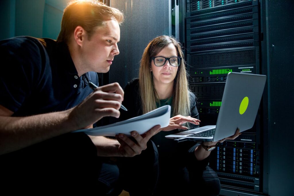Two techs in a data center working with a laptop and another device and looking very serious.