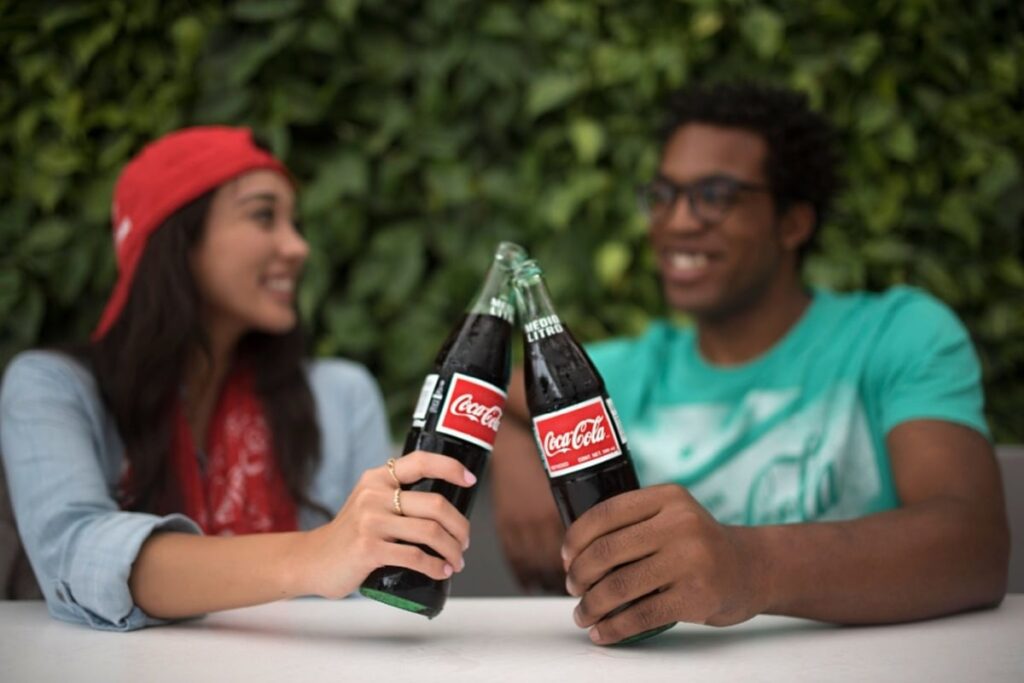 Two friends toast with glass Coca-Cola bottles against a leafy green backdrop.
