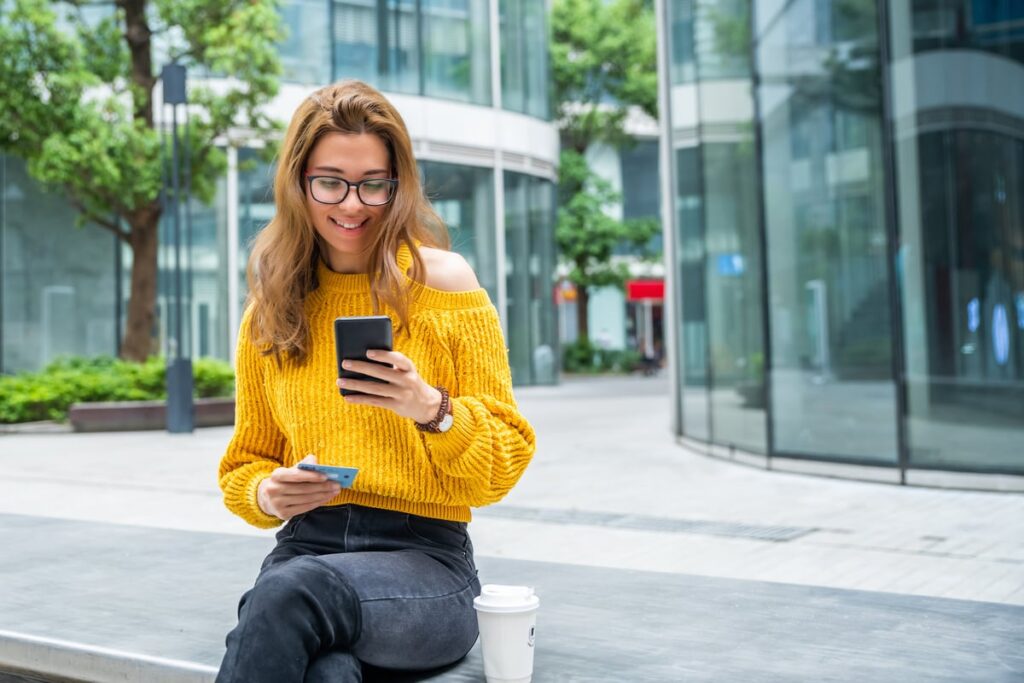 Person with a phone and a credit card is sitting on a stone bench within walking distance of financial institutions in a city.