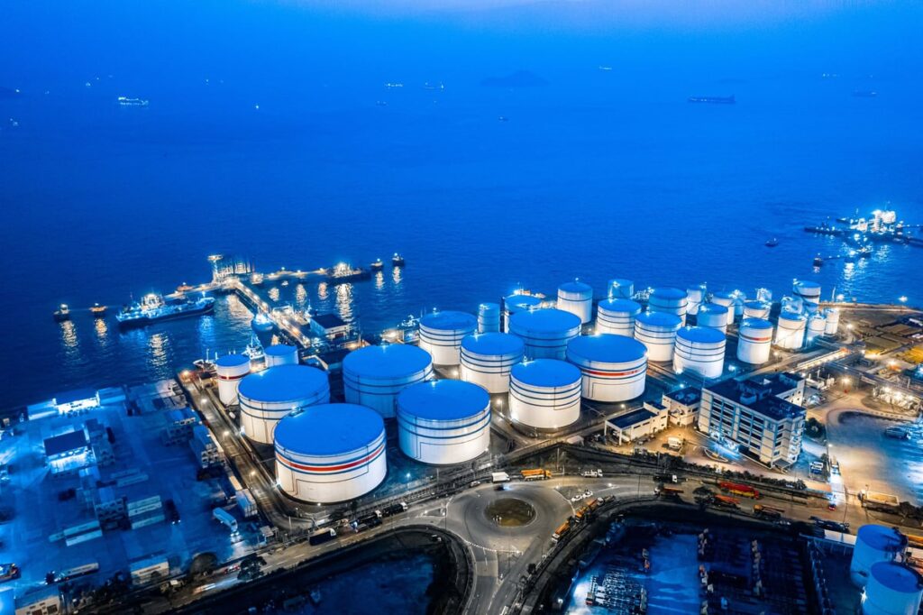 An aerial view of an oil and chemical storage terminal with large cylindrical storage tanks situated along a coastal harbor.